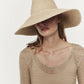 Woman wearing a large Janessa Leone Tinsley hat and a beige mesh top, posing against a white background. Her face is partially obscured by the hat.