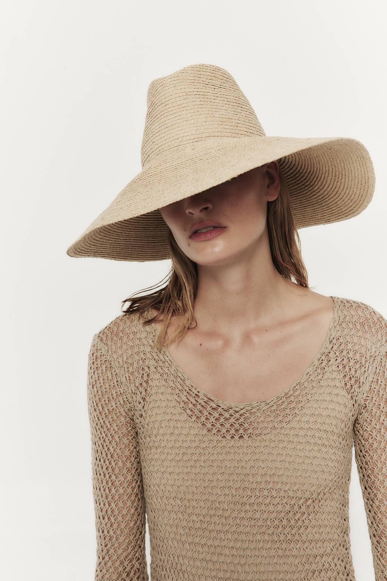 Woman wearing a large Janessa Leone Tinsley hat and a beige mesh top, posing against a white background. Her face is partially obscured by the hat.