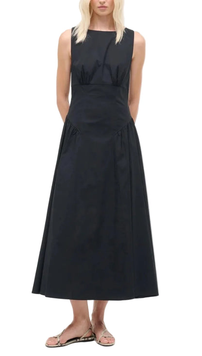 A woman wearing the Staud Gwen Dress, a sleeveless black ankle-length A-line dress with a fitted waist, structured pleats, and flared skirt, styled with flat sandals.