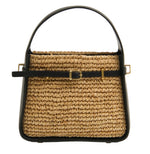 The Simkhai Cleo Mini Raffia Bucket Bag features a woven straw design with black leather handle, trim, and belt-style strap with a gold buckle and hardware, shown against a white background.