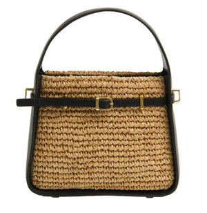 The Simkhai Cleo Mini Raffia Bucket Bag features a woven straw design with black leather handle, trim, and belt-style strap with a gold buckle and hardware, shown against a white background.