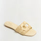 The Simkhai Monogram Raffia Flat Slide features a beige woven design with a wide frayed strap and square buckle detail—sleek flats by Simkhai, perfect for elevating your casual wardrobe, shown on a plain light background.