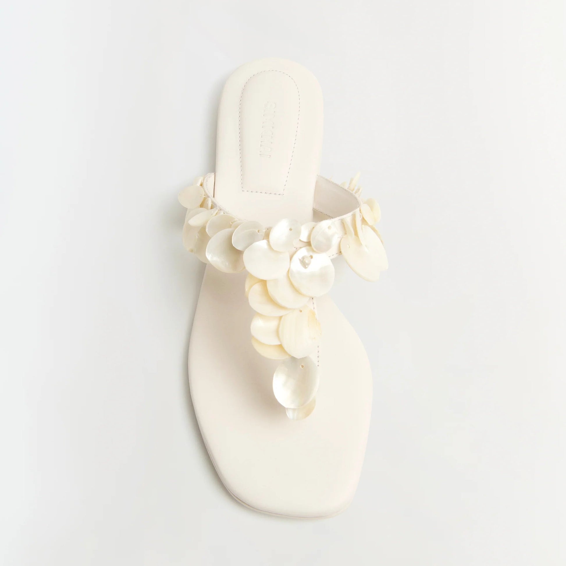The Simkhai Elin Natural Shell Beaded Thong Sandal is a white flat sandal featuring an open toe and a thong silhouette, adorned with layered, round shell-like beaded embellishments on the strap.