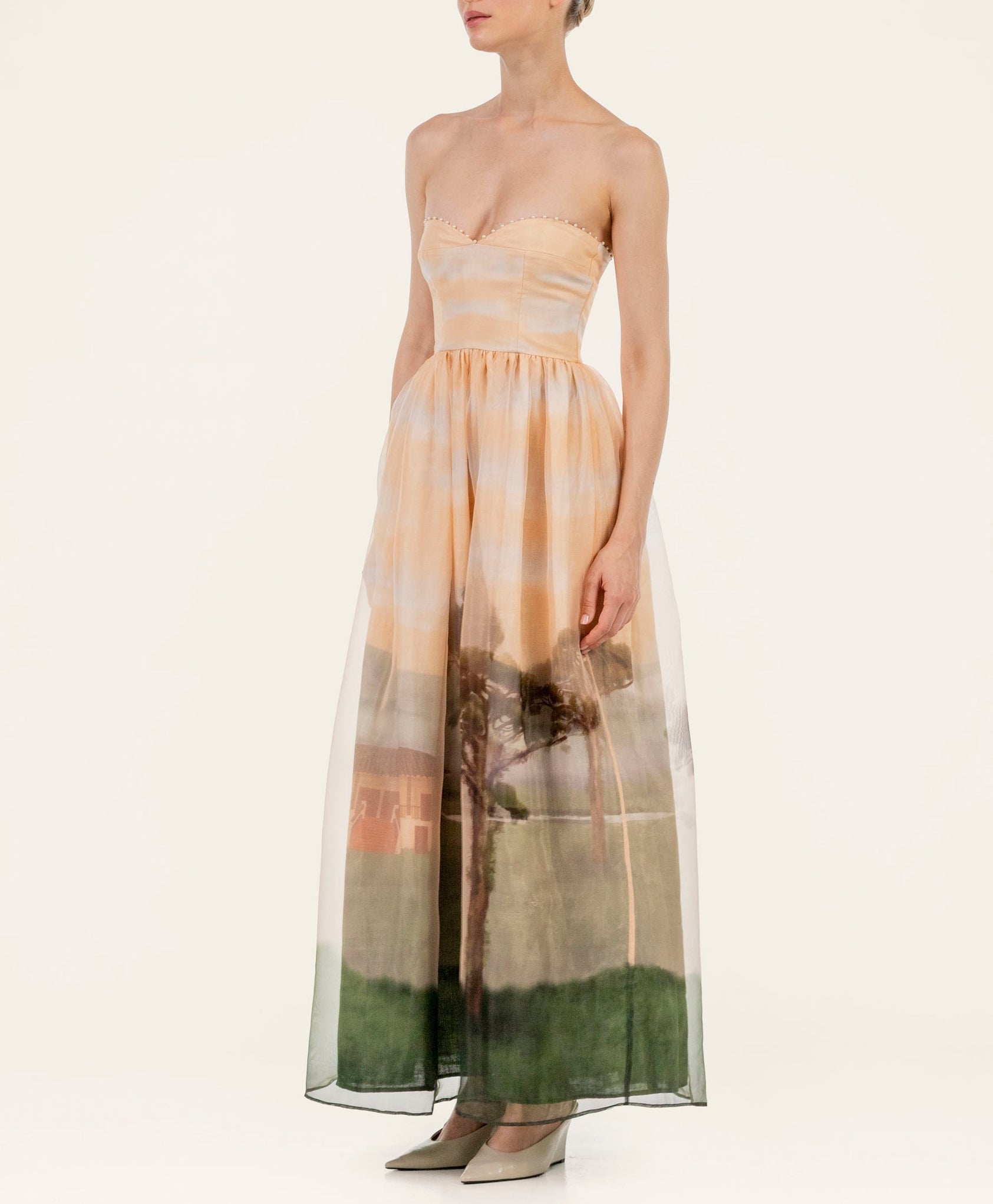 A woman wears the Tonia Dress by Agua By Agua Bendita, featuring a strapless fitted bodice and landscape print skirt—ideal for special occasions—as she stands against a plain background in pointed-toe shoes.
