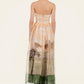 A woman faces away, wearing the Tonia Dress by Agua By Agua Bendita—a strapless occasion dress with a landscape print in soft peach and green, showcasing trees and grass, ideal for formal events.