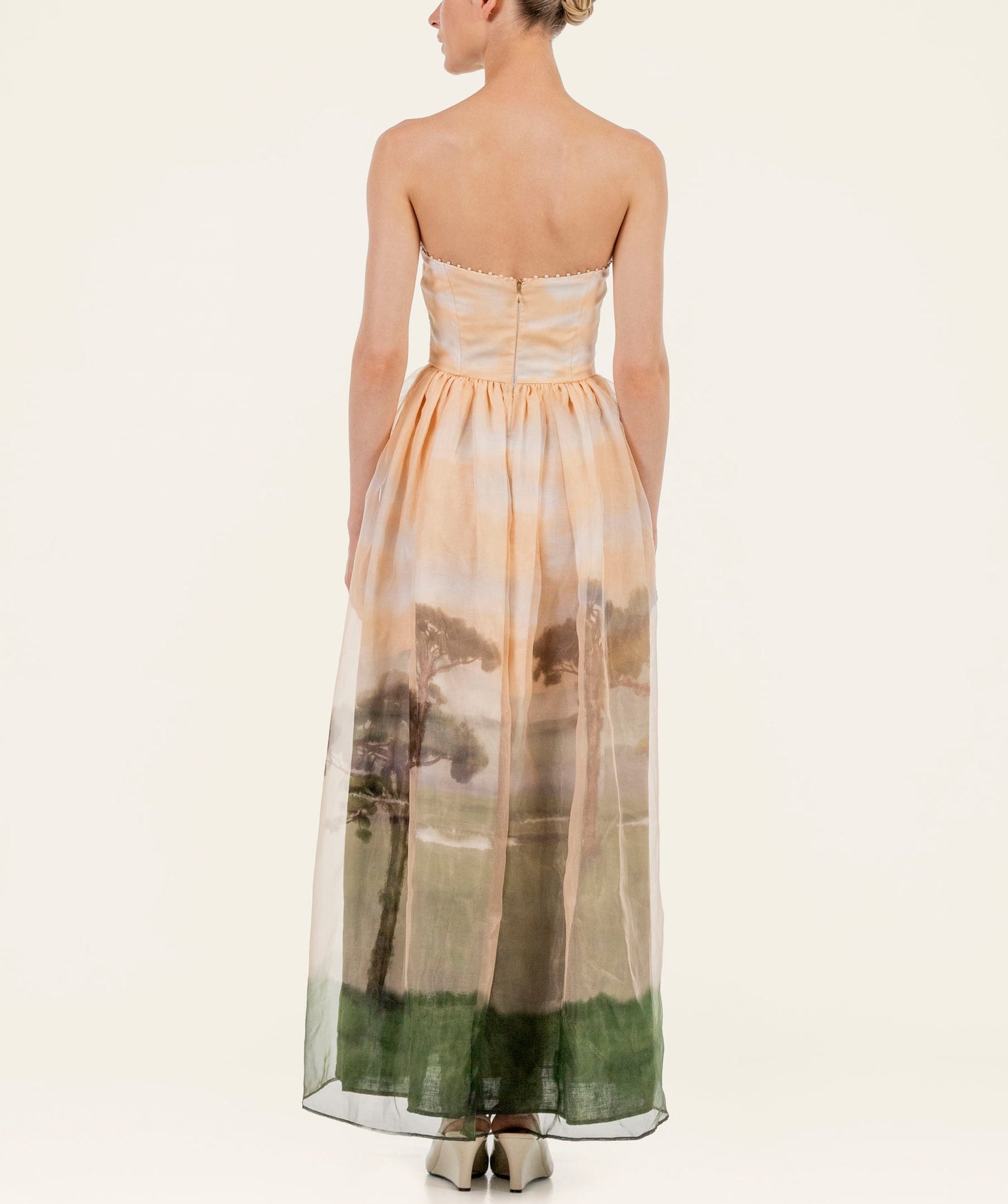 A woman faces away, wearing the Tonia Dress by Agua By Agua Bendita—a strapless occasion dress with a landscape print in soft peach and green, showcasing trees and grass, ideal for formal events.