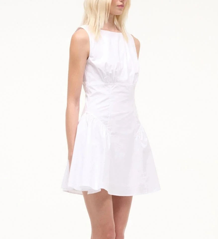 A woman stands against a plain light background wearing the Staud Gwen Mini Dress, a sleeveless white poplin A-line dress with structured pleats.