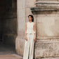 A woman in a Lela Rose Stretch Cotton Halter Top and wide-leg white trousers stands by a stone pillar in an outdoor urban setting.