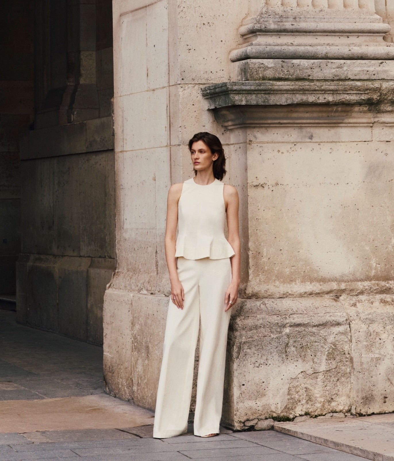 A woman in a Lela Rose Stretch Cotton Halter Top and wide-leg white trousers stands by a stone pillar in an outdoor urban setting.