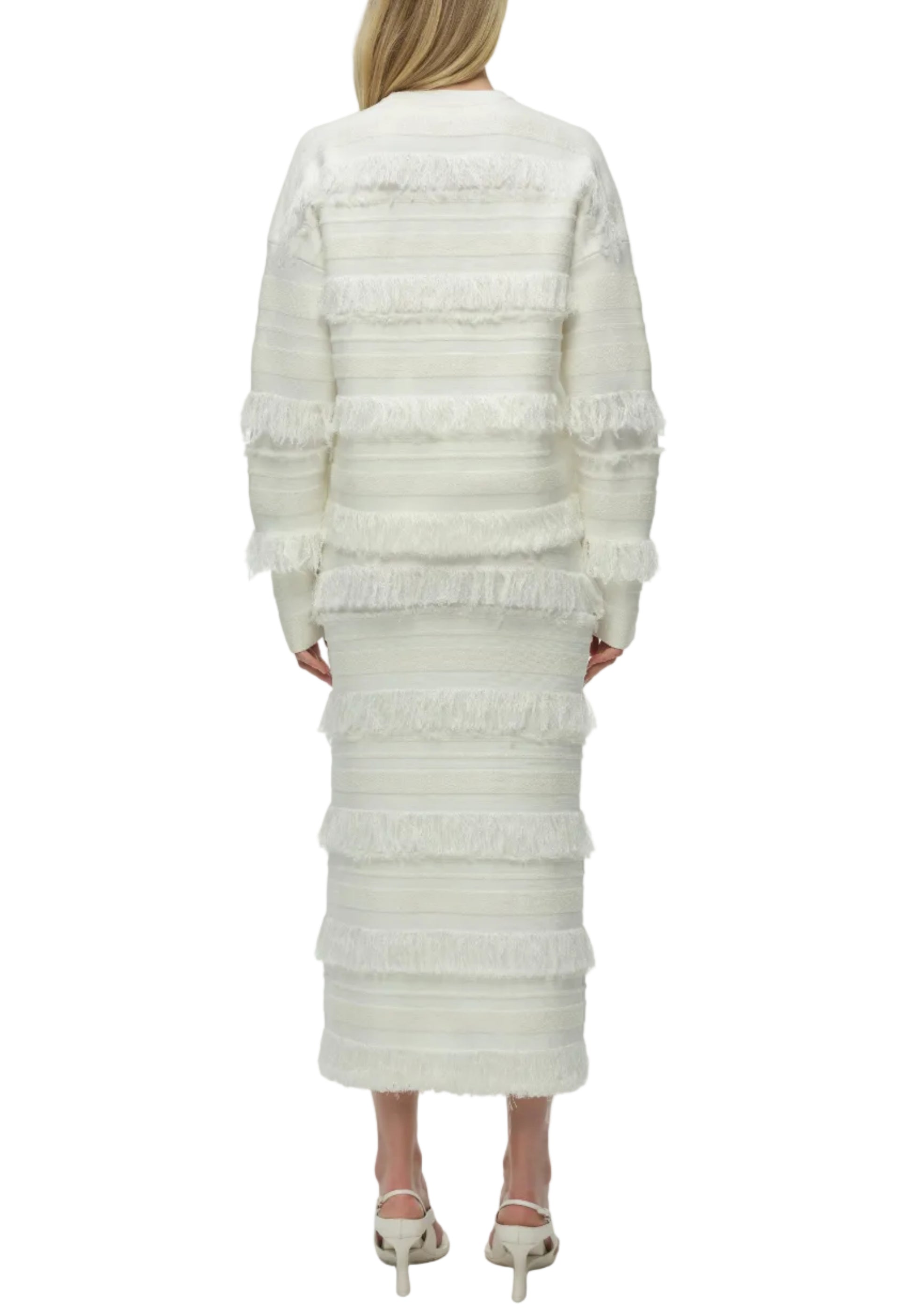 A woman faces away, wearing the Simkhai Carrick Crewneck Sweater—a white, long-sleeved knit top with fringe and horizontal stripes—paired with a matching midi skirt and white heeled sandals.