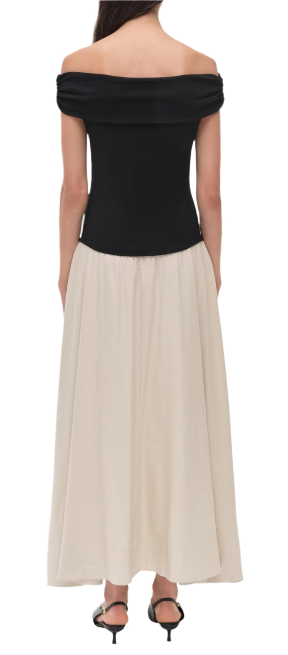 A woman stands facing away, wearing the Simkhai Adaline Off Shoulder Midi Dress with a black sculpted bodice and beige skirt, styled with black heeled sandals.