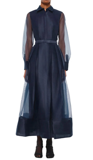 A person stands against a plain background, elegantly dressed in the Simkhai Montgomery Dress, a sheer navy midi-length creation featuring a button-up collar and belt, paired with black heels.