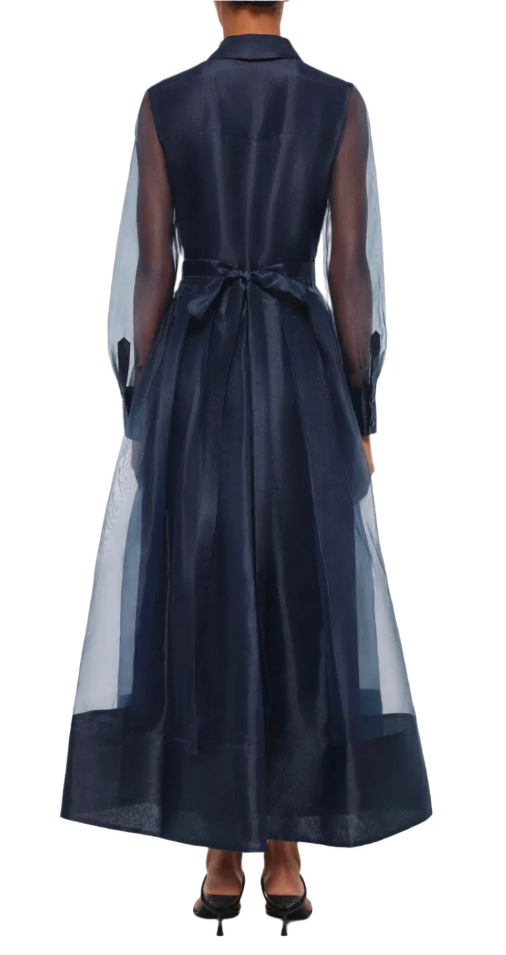 A person wearing the Simkhai Montgomery Dress, featuring a sheer long-sleeved navy design with a bow at the waist, is viewed from the back, highlighting its elegant midi-length style.