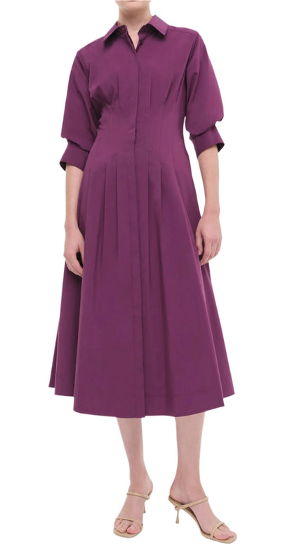 A person wearing the Simkhai Signature Jazz Dress, a purple, knee-length midi dress with elbow-length sleeves and elegant pintuck detailing, paired with beige heeled sandals from Simkhai.