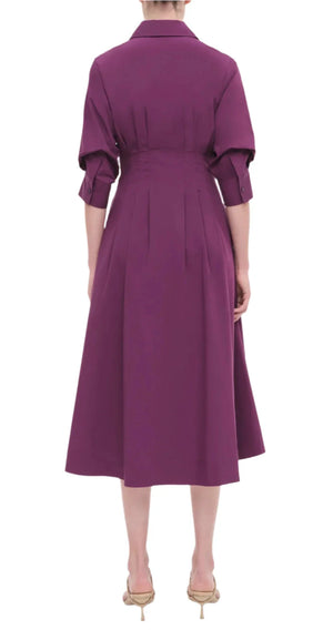 A person wearing the Simkhai Signature Jazz Dress in purple, with sleeves rolled up, is seen from the back, standing gracefully on beige heels. The tailored bodice and elegant pintuck detailing of the dress contribute a touch of sophistication to the ensemble.