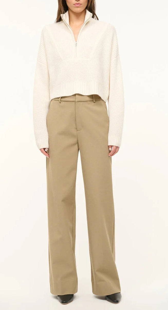 A person wears the Staud Cropped Hampton Cashmere Sweater in cream chunky knit, paired with high-waisted beige wide-leg trousers and black shoes, standing against a plain background.