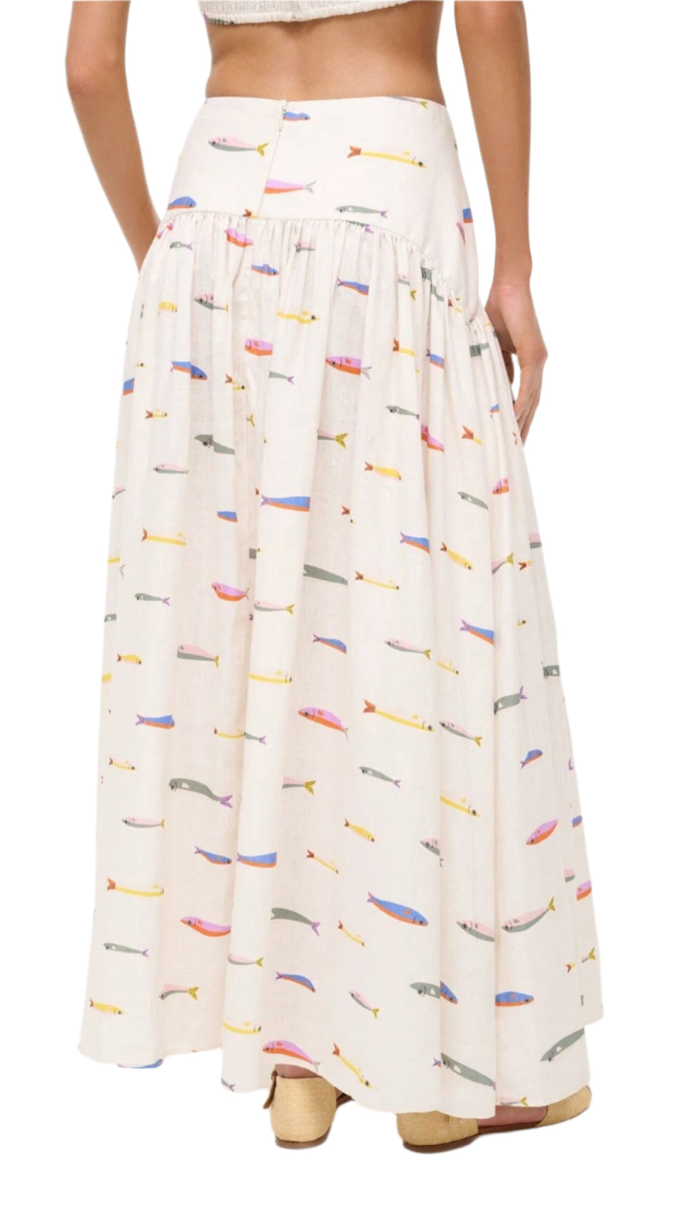 A woman stands with her back to the camera wearing the Staud Procida Linen Skirt, a high-waisted, ankle-length cream maxi skirt featuring a colorful sardines print.