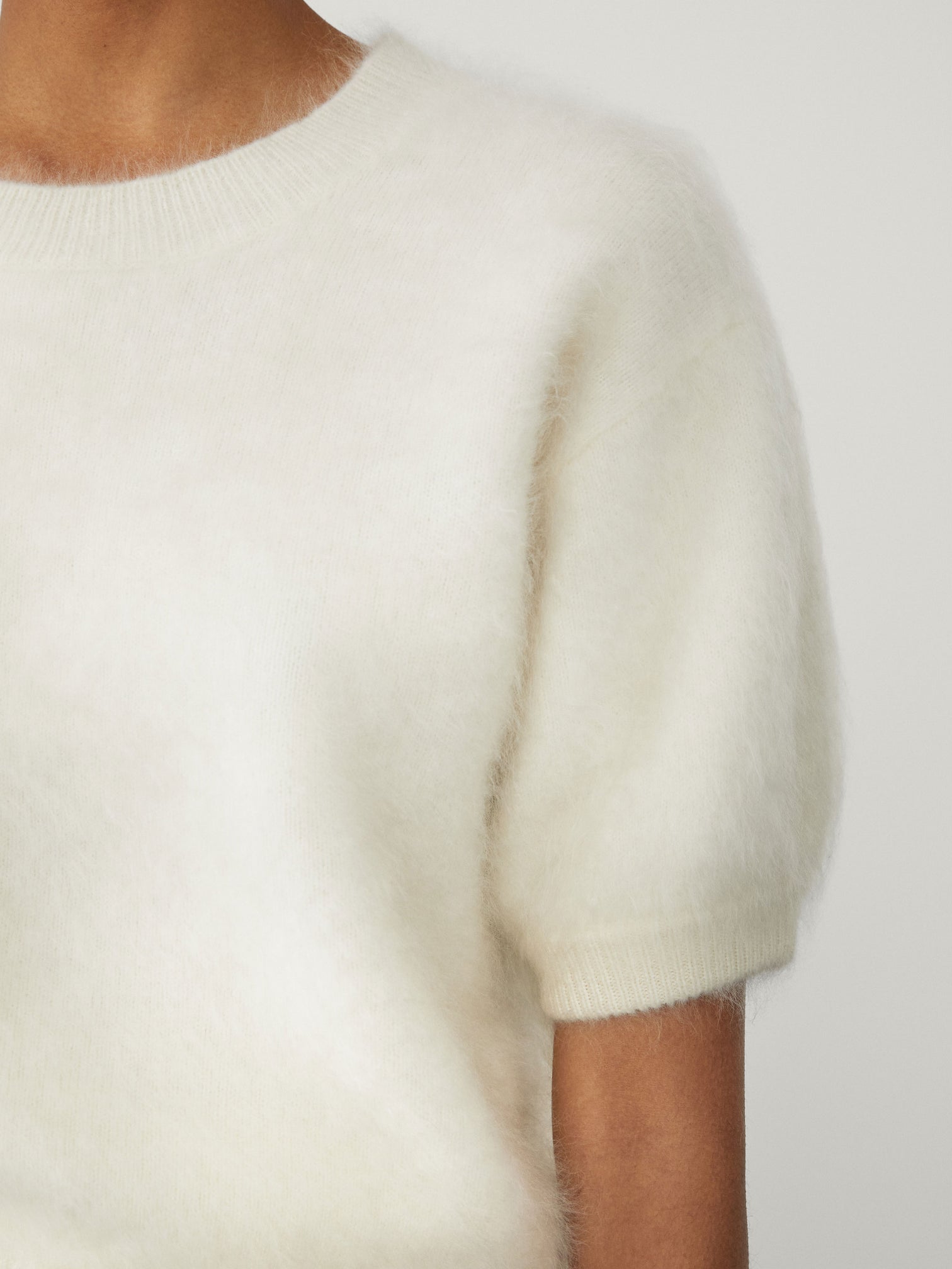 Close-up of a person wearing the Lisa Yang Juniper Brushed Cashmere Sweater, a short-sleeved, white crewneck, set against a plain background.