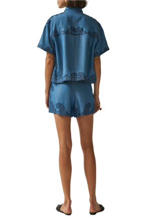 A person stands barefoot, seen from the back on a white background, wearing Carolina K’s Pina Shirt—a blue short-sleeve top with wave-like patterns and subtle embroidery—paired with matching shorts.
