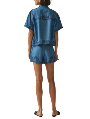 A woman stands with her back to the camera, wearing Carolina K's Roma Shorts and a matching blue Tencel denim short-sleeve shirt with black wave patterns, paired with black sandals.