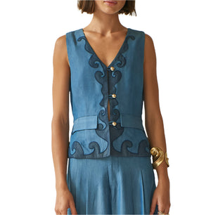 A woman wears the Carolina K Rebekka Vest, a sleeveless denim piece with front button closure and scalloped trim, paired with matching blue pleated pants and accessorized with a gold bracelet.