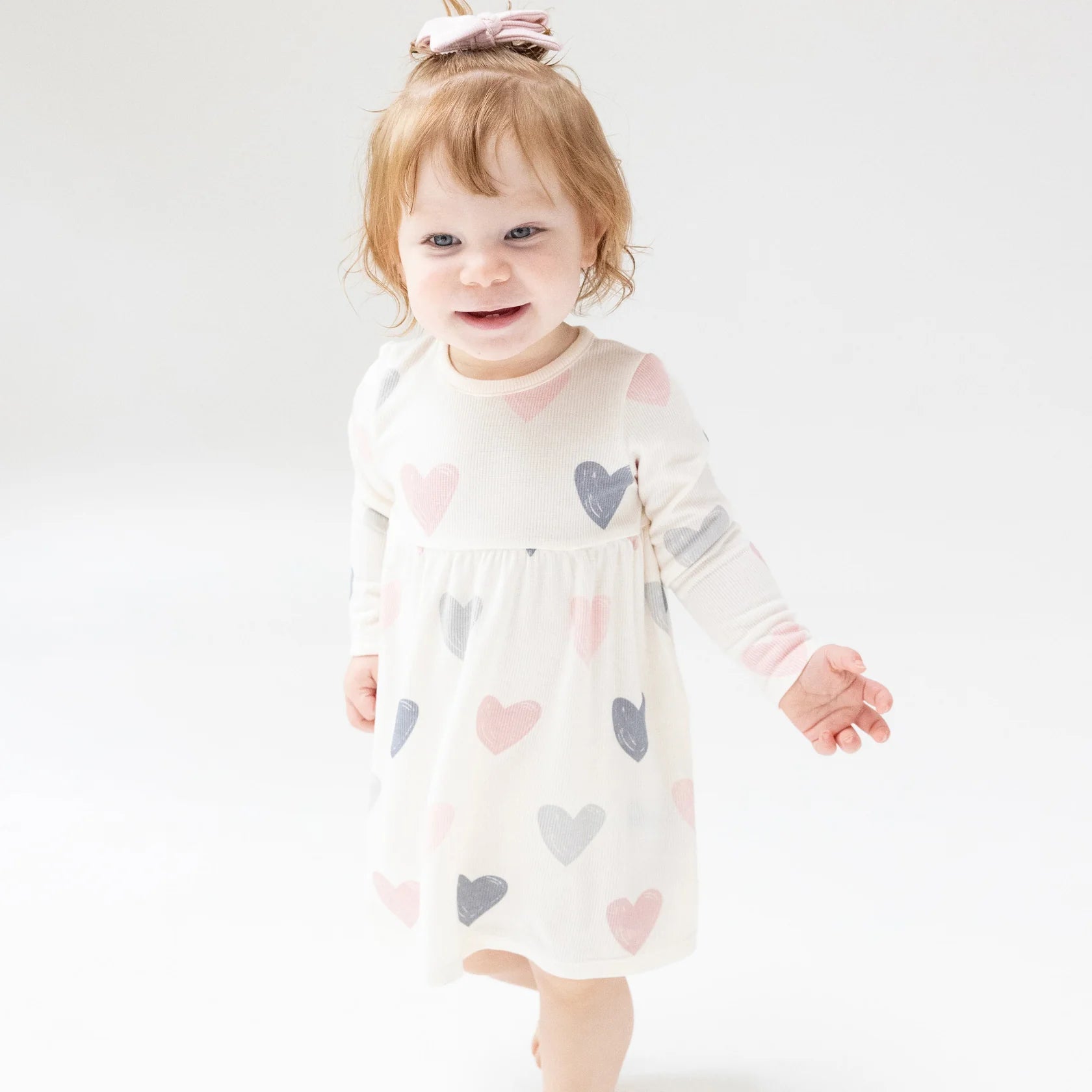 A toddler with light red hair smiles and looks to the side while wearing the cozy Angel Dear AD Modal Rib Simple Dress + Bloomer with pastel heart prints, standing against a light background.