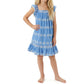A young girl with long blonde hair stands barefoot against a white background, wearing the Mer Girls’ Mini Eva Dress in blue, featuring knee-length styling, white patterns, and flutter sleeves.