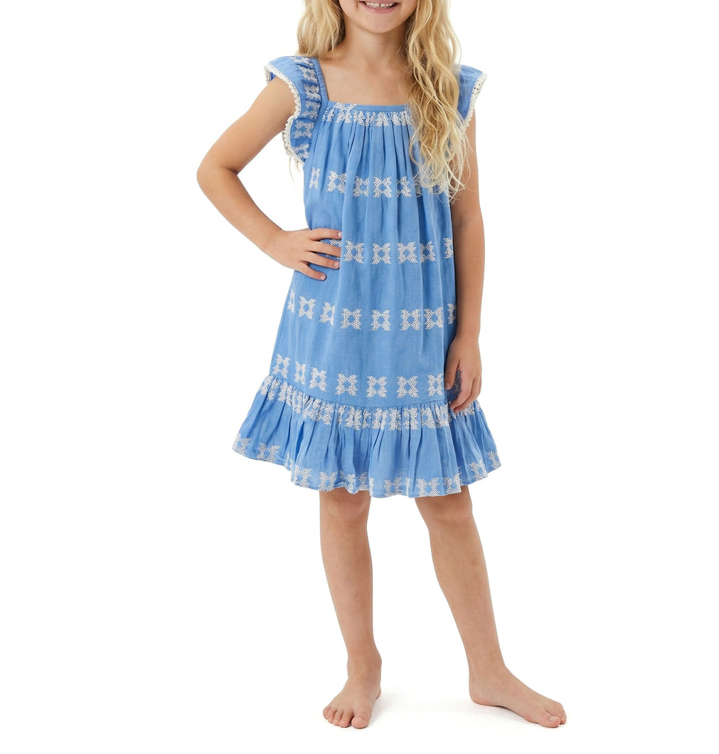A young girl with long blonde hair stands barefoot against a white background, wearing the Mer Girls’ Mini Eva Dress in blue, featuring knee-length styling, white patterns, and flutter sleeves.