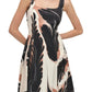 A person is wearing the Simkhai Paola Midi Dress, featuring a sleeveless, square-neck design with a black, white, and peach abstract print. The flattering silhouette is complemented by the person's hands in the dress's pockets.