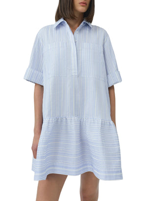 A woman wears the Simkhai Cris Short Sleeve Shirt Dress featuring blue stripes, a collared neckline, loose fit, and ruffled hem.