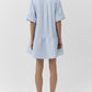 A woman stands with her back to the camera, wearing the Simkhai Cris Short Sleeve Shirt Dress in loose blue stripes and black slip-on shoes.