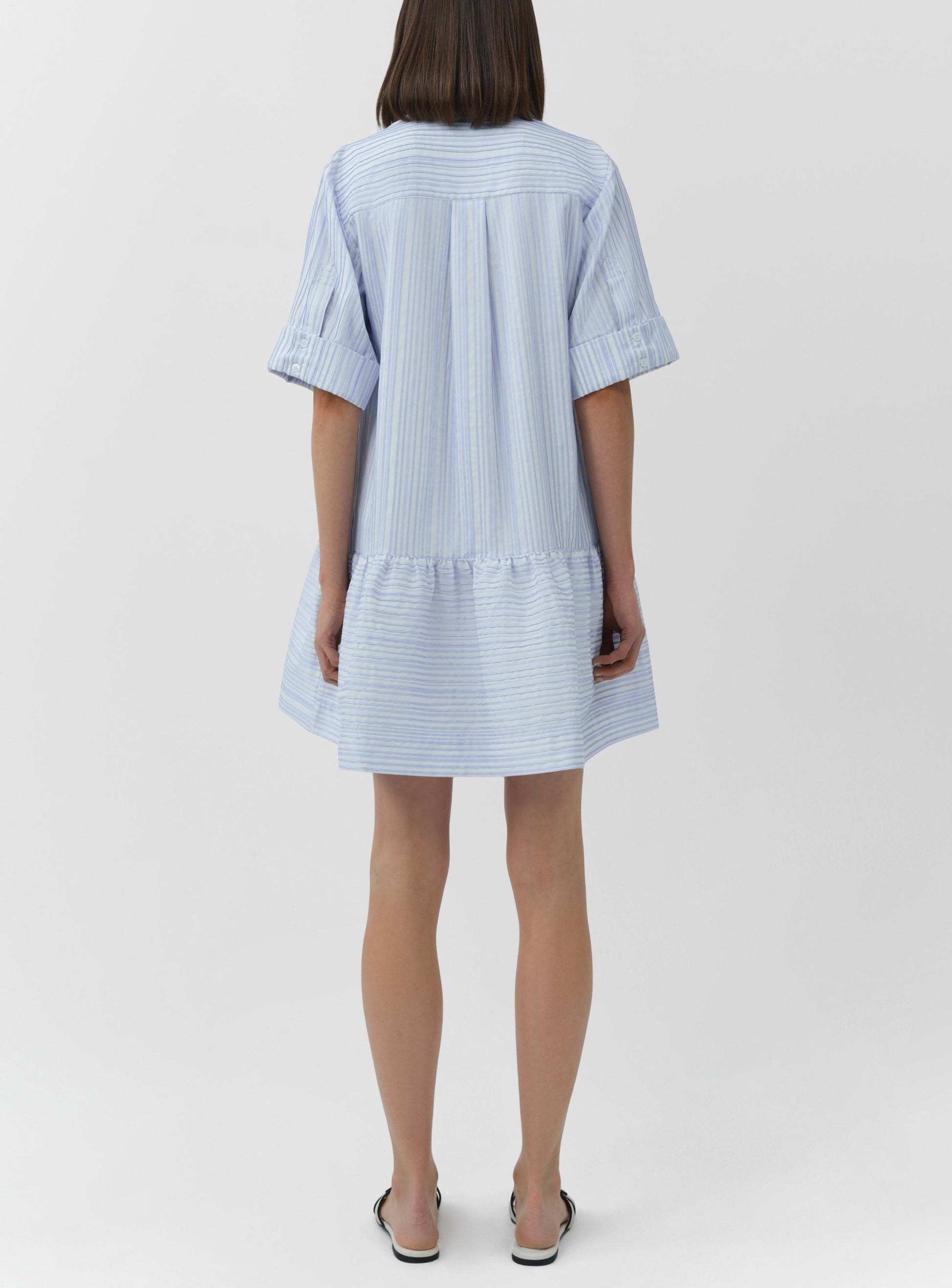 A woman stands with her back to the camera, wearing the Simkhai Cris Short Sleeve Shirt Dress in loose blue stripes and black slip-on shoes.