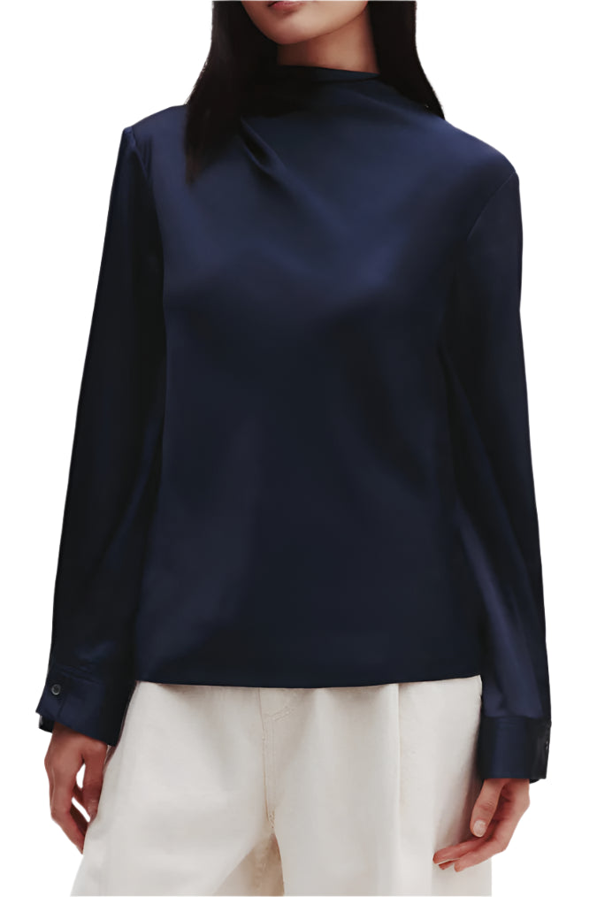 Sporting the TWP Phoebe Top, a dark long-sleeve mock neck, and light pants, the model stands against a plain background, creating an elongated silhouette.