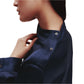 A person is shown in a profile view wearing the TWP Phoebe Top, a sleek navy button-up shirt with visible collar and sleeve buttons. The elongated silhouette contributes to an understated elegance.