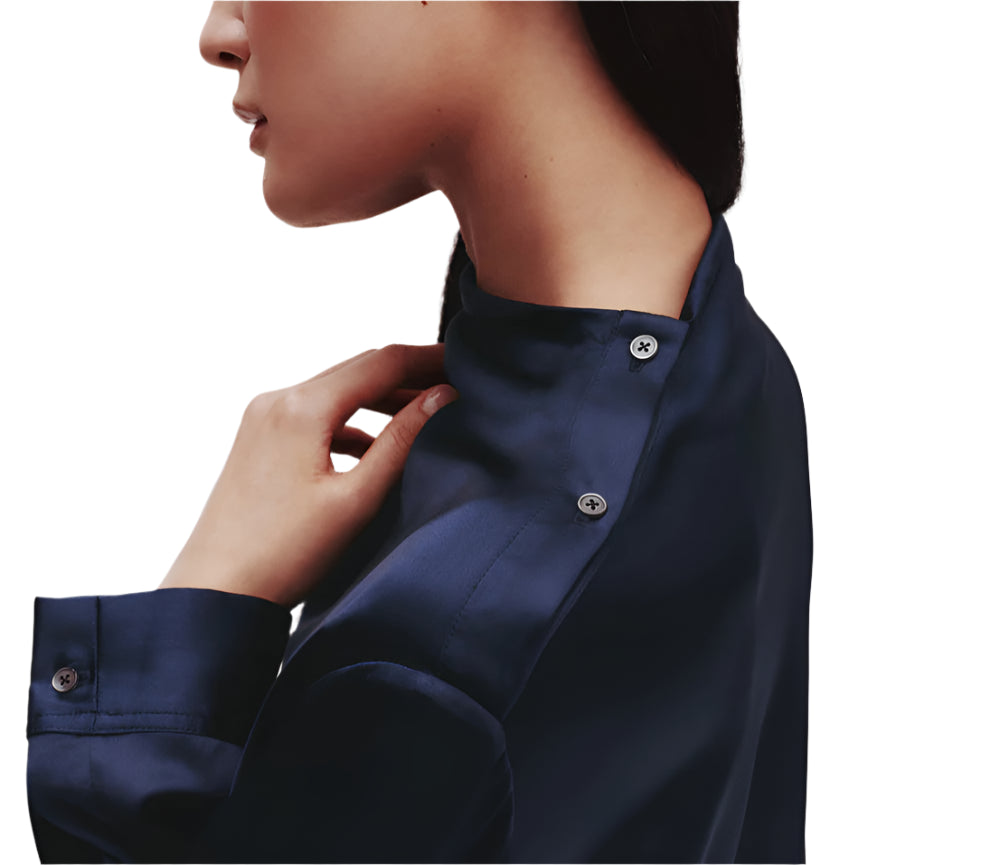 A person is shown in a profile view wearing the TWP Phoebe Top, a sleek navy button-up shirt with visible collar and sleeve buttons. The elongated silhouette contributes to an understated elegance.