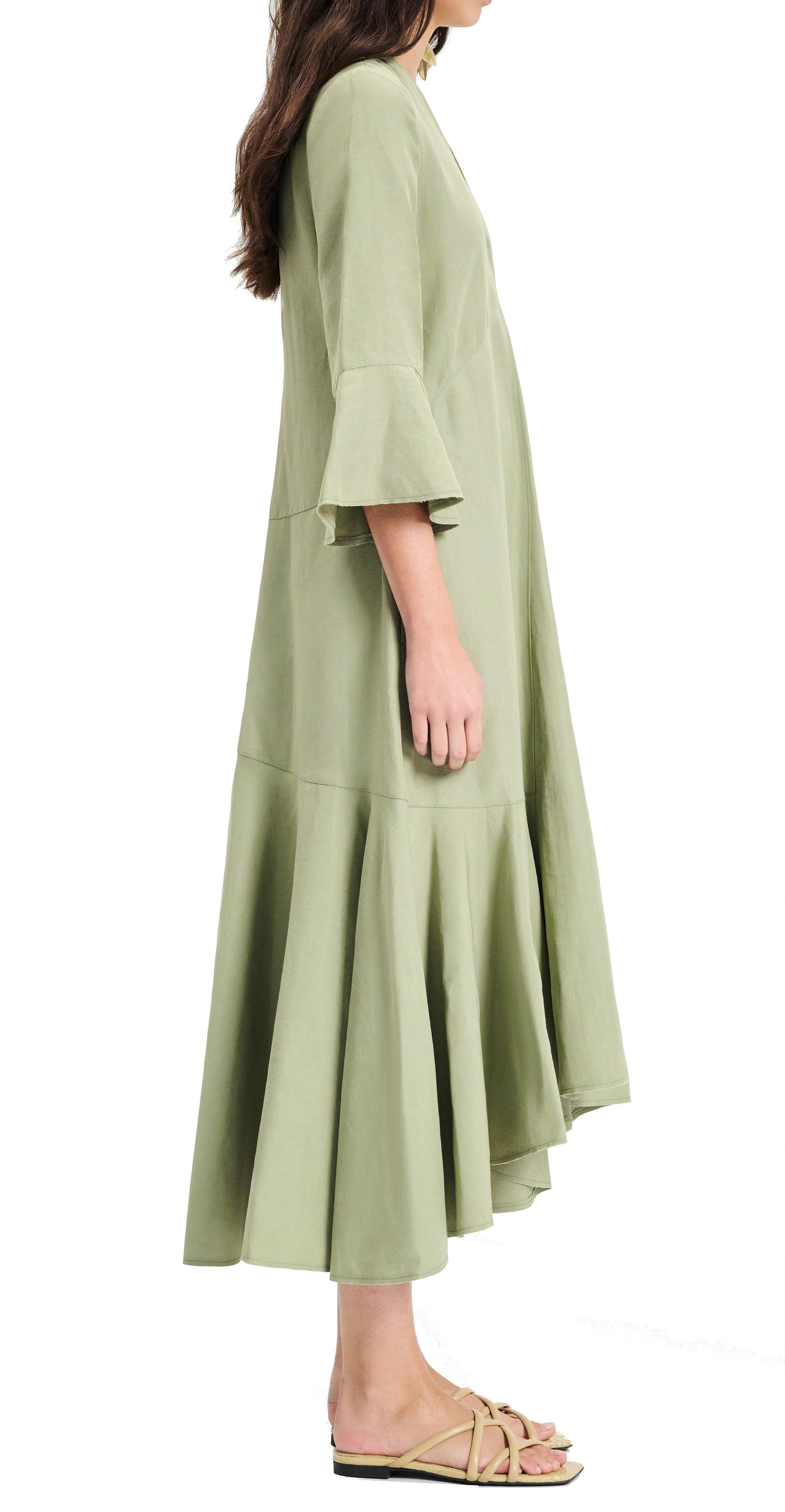 A person is standing sideways wearing the Dorothee Schumacher Summer Cruise Maxi Dress, a light green linen blend dress with flared sleeves and hem, paired with beige sandals.