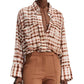 A person wearing the Dorothee Schumacher Checked Statement Blouse, characterized by its oversized style with wide sleeves in a brown and white plaid pattern, tucked partly into high-waisted brown pants.