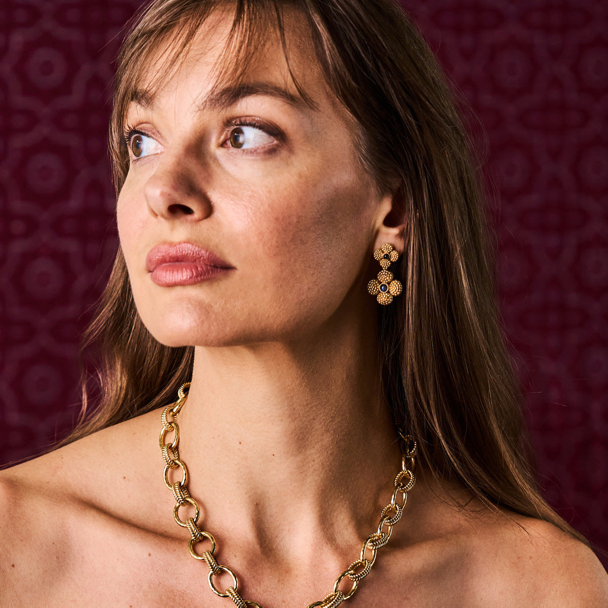A woman with light brown hair looks to the side, wearing Juliska Capucine De Wulf Berry Clover Convertible Duo Earrings and a chunky gold chain necklace against a patterned maroon background.