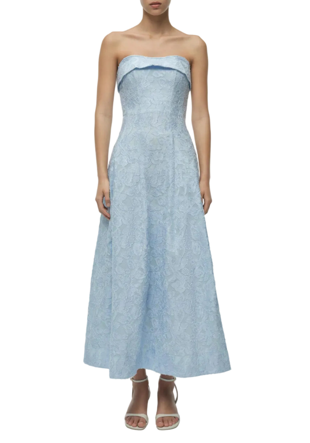 A woman wears the Simkhai Valeria Jacquard Dress—strapless, ankle-length, with a fitted bodice and flared skirt—in light blue lace, styled with white heeled sandals.