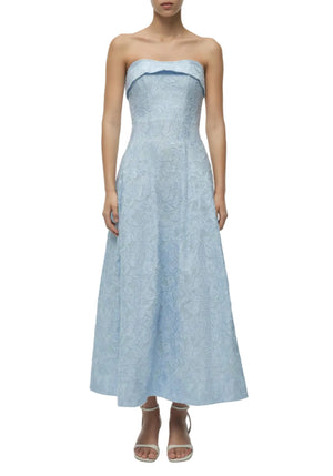 A woman wears the Simkhai Valeria Jacquard Dress—strapless, ankle-length, with a fitted bodice and flared skirt—in light blue lace, styled with white heeled sandals.