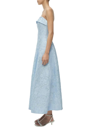A woman in the Simkhai Valeria Jacquard Dress, a light blue strapless organza style with ankle-length floral details, paired with white heeled sandals, is shown from the side against a white background.