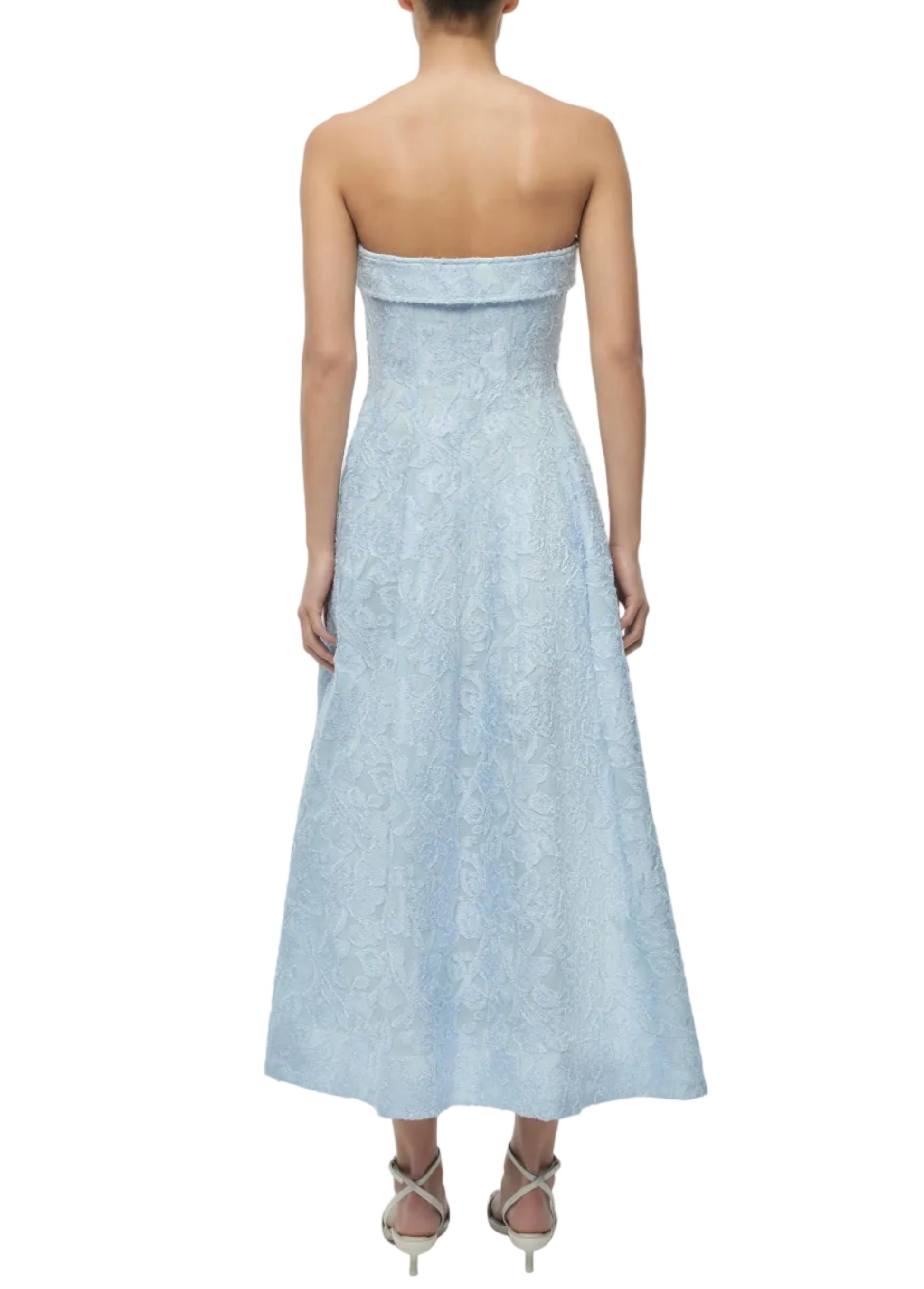 A woman, seen from the back, wears the Simkhai Valeria Jacquard Dress in light blue with floral patterns. She stands elegantly in strappy heeled sandals.
