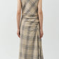 A person faces away, wearing the Simkhai Burke Draped Midi Dress in beige plaid with heeled sandals, highlighting its chic draped style against a plain light background.