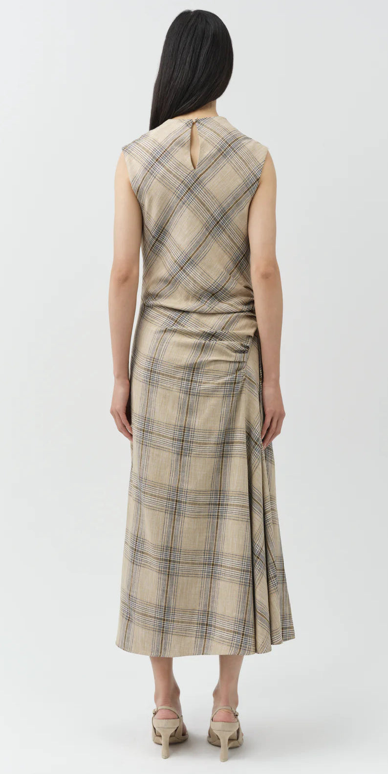 A person faces away, wearing the Simkhai Burke Draped Midi Dress in beige plaid with heeled sandals, highlighting its chic draped style against a plain light background.