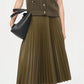 A person wears the Simkhai Perrie Vest—a sleeveless cable-knit V-neck—paired with an olive green pleated midi skirt, black knee-high boots, and a black handbag, standing against a plain background.