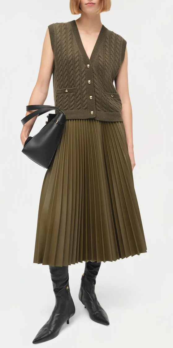 A person wears the Simkhai Perrie Vest—a sleeveless cable-knit V-neck—paired with an olive green pleated midi skirt, black knee-high boots, and a black handbag, standing against a plain background.