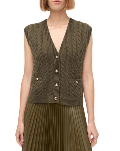 A woman wears the Simkhai Perrie Vest—an olive green, sleeveless, cable-knit cashmere vest with gold buttons—paired with a pleated skirt.