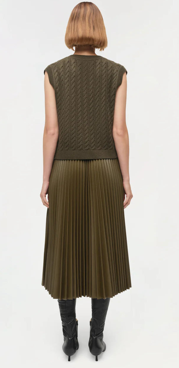 A person with short, straight hair is shown from the back wearing the Simkhai Perrie Vest, a sleeveless dark green cable knit top with a V-neckline, paired with a pleated skirt and black knee-high boots.
