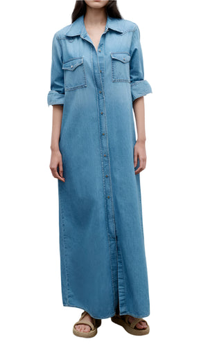 A woman wears the TWP Cowboy Maxi Dress in vintage wash cotton denim, featuring rolled-up sleeves, front pockets, and western-inspired details, paired with sandals.