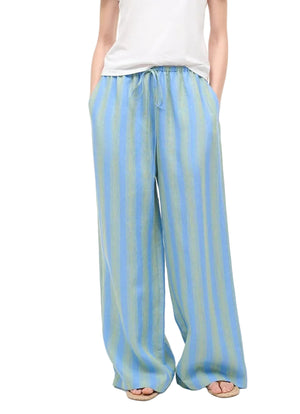 A person wears Staud's Mateo Linen Pant—loose-fitting, blue and green vertical stripes, drawstring waist, relaxed leg—with hands in pockets, styled with a white top and open-toed sandals.